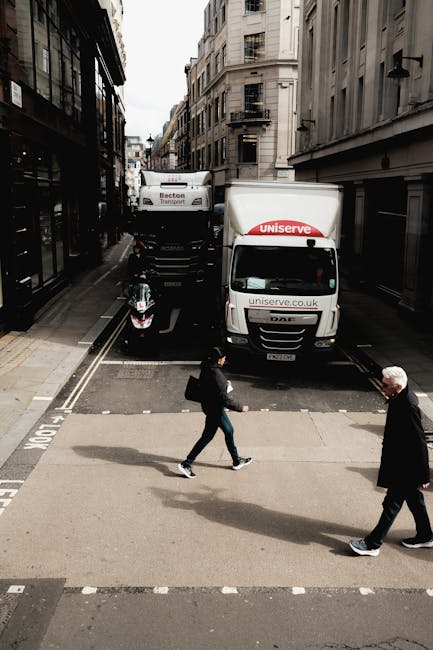 A street scene showing a moving van and a delivery truck parked closely together on a narrow urban road with tall buildings on either side. The moving van, which is part of a home relocation service provided by Man and Van Stepney, is situated in the background with its rear doors open, indicating active loading or unloading. A motorbike is parked beside the vehicles. In the foreground, a man and a woman are crossing the street at a pedestrian crosswalk; the woman is carrying a bag and walking quickly, while the older man is walking with his hands in his pockets. The scene is lit by natural daylight, showing urban infrastructure such as fire escapes and retail signage, emphasizing a busy city environment where furniture transport and packing and moving are taking place as part of a professional removals process.