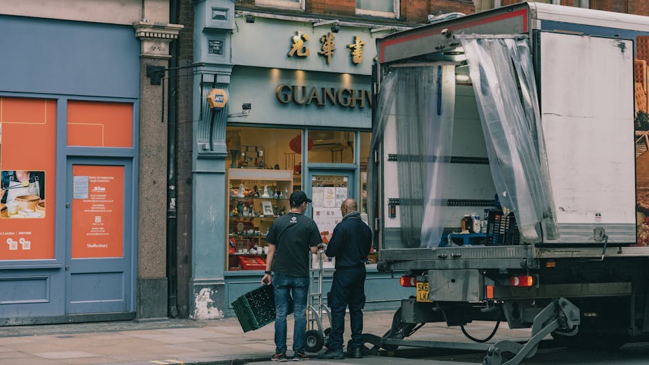 Two men, one holding a portable hand truck and the other carrying a small wheeled cart, stand outside the storefront of Guangh Chinese restaurant on a city street during daytime. The restaurant's facade features light blue walls with gold Chinese characters and English signage, and a display window showcasing various items, possibly menus or promotional materials. A large moving truck with an open side panel and transparent plastic curtain is parked nearby, indicating active loading or unloading activities associated with house removals or furniture transport. The truck is positioned on the pavement, close to the restaurant entrance, with visible equipment such as straps and blankets, suggesting a professional packing and moving process by Man and Van Stepney. The scene is well-lit with natural daylight, and the surroundings include urban elements like a blue door to the side and other signage, emphasizing the context of home relocation or moving services in the area.