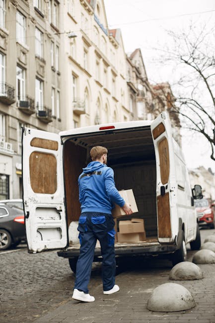 A street scene showing a moving van and a delivery truck parked closely together on a narrow urban road with tall buildings on either side. The moving van, which is part of a home relocation service provided by Man and Van Stepney, is situated in the background with its rear doors open, indicating active loading or unloading. A motorbike is parked beside the vehicles. In the foreground, a man and a woman are crossing the street at a pedestrian crosswalk; the woman is carrying a bag and walking quickly, while the older man is walking with his hands in his pockets. The scene is lit by natural daylight, showing urban infrastructure such as fire escapes and retail signage, emphasizing a busy city environment where furniture transport and packing and moving are taking place as part of a professional removals process.