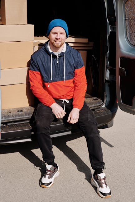 A man with a beard wearing a black cap and white polo shirt stands inside the open rear of a moving van, which is filled with neatly stacked cardboard boxes of various sizes, some wrapped in plastic o