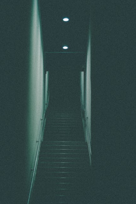 A narrow staircase inside a residential property, featuring dark wooden steps and white walls on either side. The staircase is illuminated by three ceiling-mounted recessed lights, casting soft light down the flight. The walls appear to be smooth and painted in a neutral colour. At the bottom of the stairs, there is a small landing area that leads to a doorway, which is partially visible. The environment appears quiet and tidy, with no furniture or additional objects visible on or around the staircase. In the context of house removals and packing and moving services, this image illustrates a common obstacle faced during home relocations, where narrow stairs require careful handling of furniture and boxes to prevent damage. [COMPANY_NAME], such as Man and Van Stepney, often assists with navigating such challenging staircases during furniture transport or removals in tight spaces.