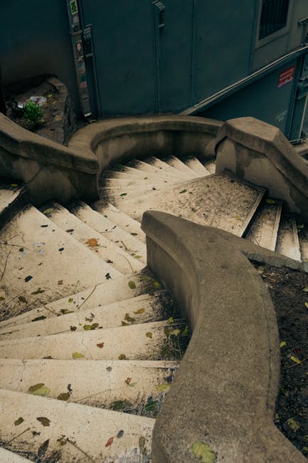 View through a dark, enclosed passageway looking out onto an exterior staircase outside a residential property. The staircase is made of weathered concrete steps leading upwards from an indoor loading area, with some steps showing signs of wear and discoloration. To the right of the stairs, there is a low concrete wall with potted plants attached, adding greenery to the outdoor space. On the left, a portion of a metal railing can be seen, supporting the stairs. At the top of the steps, a small landing is visible, surrounded by a brick and wooden fence, along with some greenery including shrubs and a tree with autumn-colored leaves. The scene captures a typical home relocation or furniture transport scenario, with the stairway presenting challenges for moving furniture and packed boxes, which [COMPANY_NAME] might need to navigate during a house removal in the area near Stepney. Natural lighting highlights the outdoor environment and the structural details of the staircase, emphasizing the importance of careful planning in narrow stairway furniture transport or packing and moving operations.
