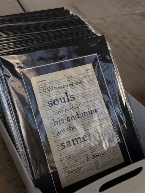 A close-up view of a white plastic crate containing framed and unframed documents, with one visible framed print featuring an excerpt from Heights Wurttemberg, displaying a quote about souls being mad