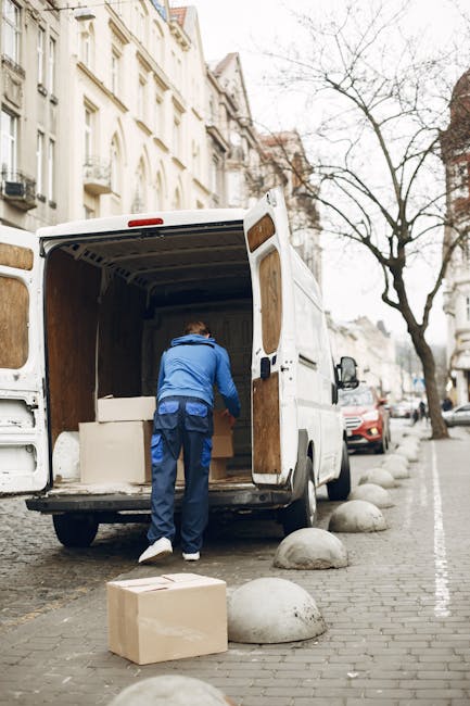 A city street scene showing a silver moving van parked on the pavement outside multi-storey buildings with mixed brick and stone facades, some with large windows and decorative architectural details. The van is positioned near the sidewalk, ready for loading or unloading furniture and boxes as part of a home relocation process. The adjacency of the vehicles and buildings indicates a busy urban environment suitable for house removals and furniture transport services conducted by Man and Van Stepney. The street is bordered by metal bollards, and the surrounding structures include historic and modern architectural elements, suggesting a central or residential area in Stepney. This scenario reflects an ongoing house removal or moving operation where packing materials, such as cardboard boxes or wrapped furniture, might be in proximity but are not visible in the image. The natural daylight and open street atmosphere emphasize the logistics involved in local or urgent removals within an urban setting.