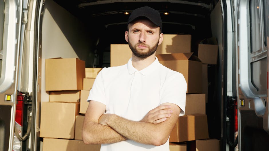 A young man with a beard, wearing a black cap and a white polo shirt, stands with arms crossed inside the open rear of a delivery van. The interior of the van is filled with stacked cardboard boxes of