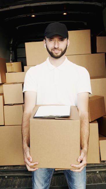 A young man with a beard, wearing a black cap and a white polo shirt, stands with arms crossed inside the open rear of a delivery van. The interior of the van is filled with stacked cardboard boxes of