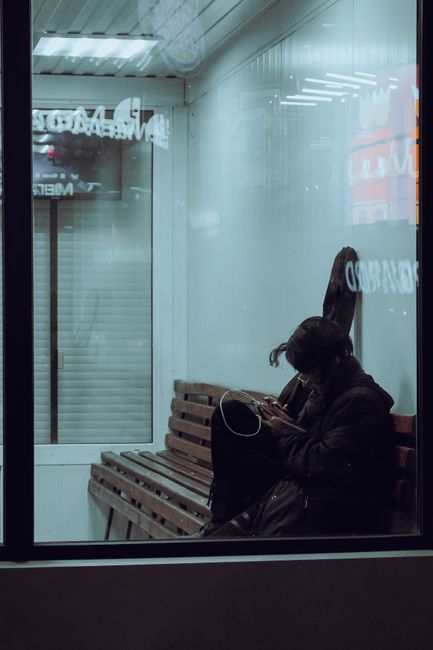 A woman sitting alone on a wooden bench inside a waiting area near a large window, with her head slightly bowed while using a smartphone. She is wearing a dark coat and has long dark hair tied back. The environment is illuminated by overhead fluorescent lighting, and outside the window, there is a reflective surface with blurred signage visible, indicating an urban setting. The scene suggests a moment of pause during a home relocation or moving process, possibly waiting for the next stage of furniture transport or packing and moving services. This setting may be associated with delays or timing issues related to late building access, as referenced in the page titled 'Late building access delays and removals timing in Stepney' on manandvanstepney.co.uk, where professional removals by Man and Van Stepney are provided.