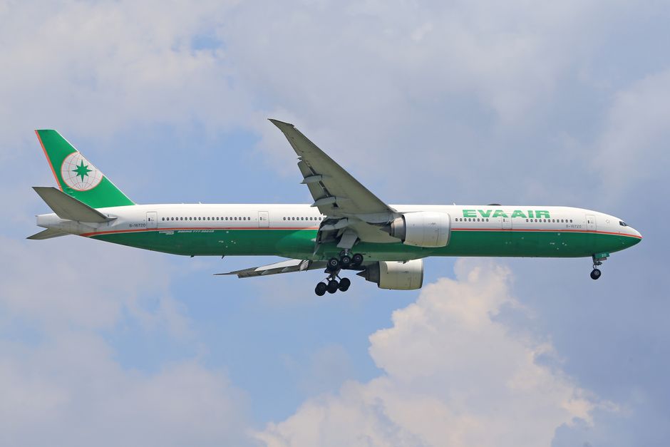 A large passenger aircraft with a green and white livery, displaying the EVA Air logo on the tail fin, is flying in the sky above a partly cloudy backdrop. The plane’s undercarriage, including wheels and landing gear, is extended as it approaches for landing or takes off. Inside the aircraft, windows are visible along the fuselage. This scene is not related to house removals or moving services, indicating a mismatch for the specified context.