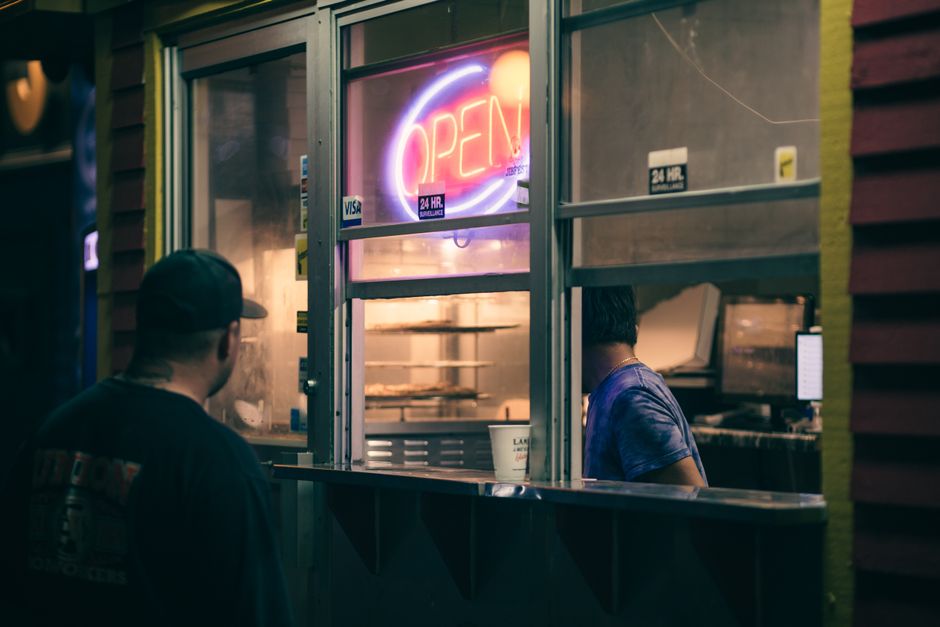 A woman sitting alone on a wooden bench inside a waiting area near a large window, with her head slightly bowed while using a smartphone. She is wearing a dark coat and has long dark hair tied back. The environment is illuminated by overhead fluorescent lighting, and outside the window, there is a reflective surface with blurred signage visible, indicating an urban setting. The scene suggests a moment of pause during a home relocation or moving process, possibly waiting for the next stage of furniture transport or packing and moving services. This setting may be associated with delays or timing issues related to late building access, as referenced in the page titled 'Late building access delays and removals timing in Stepney' on manandvanstepney.co.uk, where professional removals by Man and Van Stepney are provided.