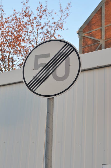 A circular traffic sign with a white background and a black diagonal stripe across it, indicating the end of a 50 miles per hour speed limit zone. The sign is mounted on a metal pole positioned in front of a corrugated white metal fence. Behind the fence, there are branches of a tree with autumn-colored leaves and a partly visible red-brick building with a sloped roof. The sky above is clear and blue. The sign's placement suggests it is located near a residential or commercial area, possibly along a street used during home relocation or furniture transport. The image is clear, with natural lighting highlighting the sign and surrounding environment, and is relevant to house removals and moving services provided by Man and Van Stepney for areas like Mile End Road flats, STEPNEY.