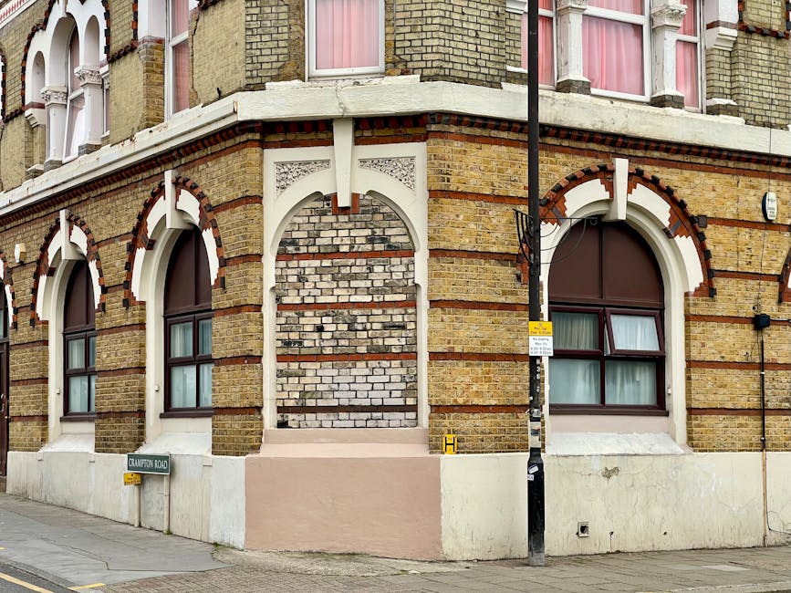 Photograph of the corner of a historic brick building on Clayton Road, featuring large arched windows with dark frames and pink curtains inside. The building has decorative white trim around the windows and arches, with red brick accents and a textured brick pattern. A black lamppost with a yellow and white bus stop sign is positioned at the street corner, overlapping part of the building façade. Below the windows, a white-painted concrete base lines the exterior wall. The scene is captured during daylight with clear weather. This image relates to house removals and furniture transportation, illustrating a typical building façade where packing and moving logistics might be coordinated by services like Man and Van Stepney during home relocations or apartment moves.