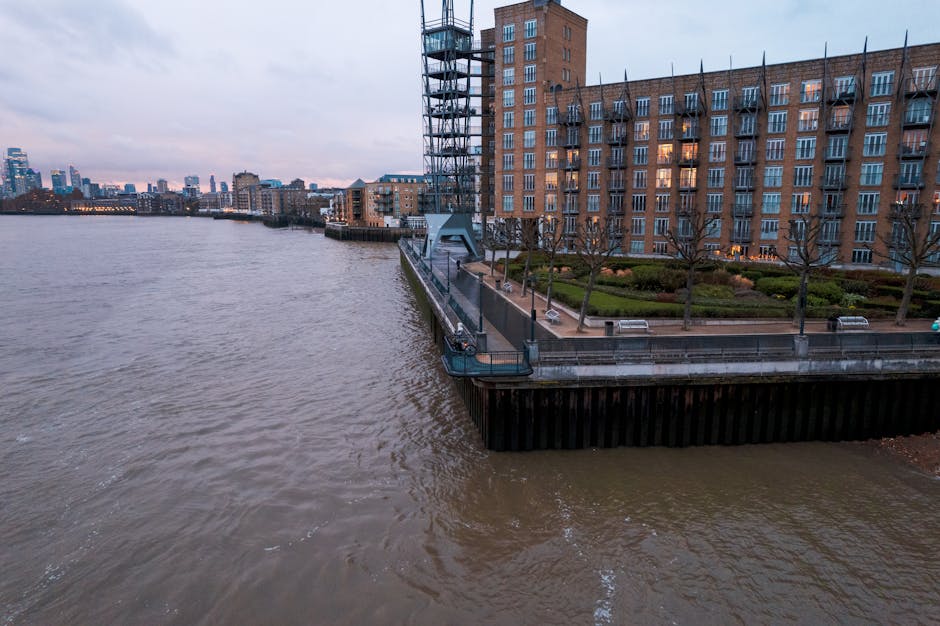 A wide view of a riverbank during dusk, featuring a modern residential building with multiple floors and numerous windows along the promenade. The building has a brick facade with a terrace area that includes a neatly landscaped garden, benches, and leafless trees. A metal security railing runs along the edge of the concrete walkway, which is adjacent to the water. A scaffolding structure is visible next to the building, indicating ongoing maintenance or development. The river appears calm with gentle ripples on the surface, reflecting the cloudy sky above, and the city skyline with high-rise buildings can be seen in the distance. The scene, captured in natural lighting, exemplifies an urban riverside environment suitable for home relocation or furniture transport activities, consistent with house removals and moving services provided by Man and Van Stepney, especially in the context of complying with Tower Hamlets council rules for furniture removals.