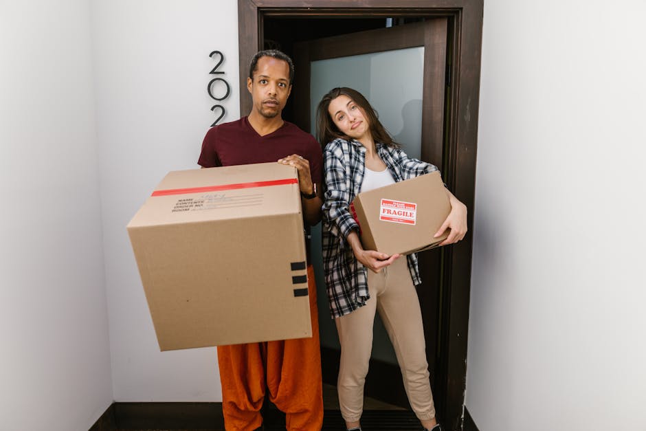 A man and a woman stand inside a narrow hallway near an open doorway, both holding cardboard boxes used for home relocation. The man, dressed in a maroon t-shirt and orange pants, holds a large box in front of him, while the woman, wearing a checkered shirt and beige pants, holds a smaller box labeled 'FRAGILE' in her arms. The wooden door frame behind them separates the interior from the exterior, and the hallway walls are painted light grey. Visible near the door is a vertical sign with the number '200', indicating the property address, and the area is illuminated by natural or artificial light. This scene depicts the loading process during furniture transport, coordinated by [COMPANY_NAME], with the individuals preparing belongings for moving or packing within a property for a house removal service, consistent with regulations for furniture moves in Tower Hamlets.