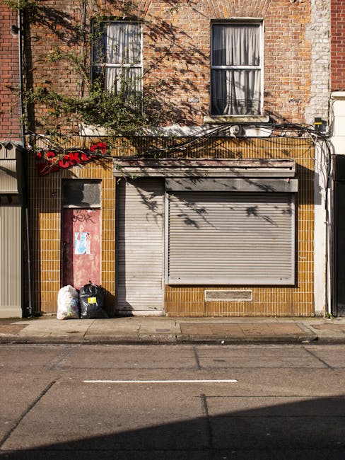 The image depicts the façade of a two-story brick building with two large, closed metal roller shutters on the ground floor, suggesting a commercial premises or storage area. To the left of the shutters, there are two black garbage bags and several clear plastic bags filled with waste or recyclables placed on the sidewalk. Above the ground floor, there are two rectangular windows with white frames and partially drawn curtains, set into the brickwork. A small amount of greenery, including a tree and some leafless branches, extends across the front of the building, casting shadows onto the façade. The street in front features a paved sidewalk and a section of asphalt with a faint white line, indicating road markings. The overall scene reflects an urban environment that could be relevant for home relocation, furniture transport, or moving logistics, with Man and Van Stepney occasionally mentioned in context of removals services.