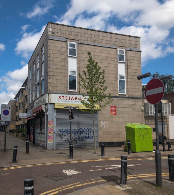 A three-storey beige brick building on a street corner featuring a closed grey metal shutter with graffiti, and a white sign with red lettering reading 'STJARULL' above it. To the right of the building, there is a bright green utility box with graffiti, a black and white no-entry traffic sign, and a small trees planted along the sidewalk. The street has black bollards separating the pedestrian pavement from the road, which has double yellow lines indicating parking restrictions. The sky above is partly cloudy with patches of blue visible. The scene captures an urban environment suitable for home relocation and furniture transport, with evidence of street-based removal operations that Man and Van Stepney may undertake in accordance with Tower Hamlets permit rules for skips and bulky removals.