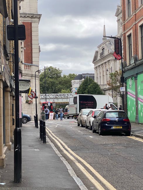 A narrow urban street scene showing a home relocation process outside a property on Tredegar Road. In the foreground, there are black bollards lining the edge of the pavement on the left, with a small sign for 'Emilia' attached to a building. Several cars are parked along the curb, including a dark-colored vehicle closest to the camera. Further down, a white moving van belonging to Man and Van Stepney is parked partially on the pavement near a doorway, with its rear doors open and a crew member visible nearby, potentially involved in the loading process. Behind the van, there are additional parked cars and pedestrians walking along the street. The background features historic-looking white stone and red brick buildings, with some greenery visible in the trees, and a cloudy sky overhead. This scene depicts a typical home packing and furniture transport logistics event, with a focus on the street-level environment involved in house removals and moving services.