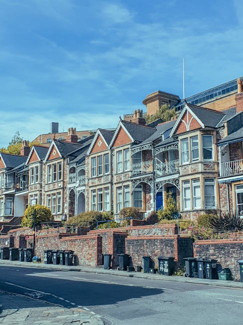 A row of Victorian-style terraced houses with ornate white ironwork balconies and decorative woodwork along the upper floors, set against a bright blue sky with some wispy clouds. The houses have brick facades, large front windows, and steeply pitched roofs. In front of the houses, there is a low brick wall with a series of small, well-maintained shrubs and trees. Along the street, black wheelie bins are lined up on the pavement, which appears clean and quiet. This image highlights typical residential architecture in the area, relevant to house removals or home relocation services provided by Man and Van Stepney, with the scene capturing the exterior environment before or after a house move.