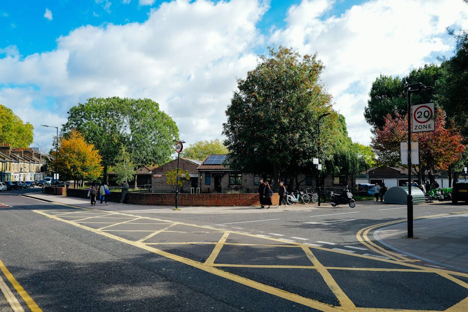 A narrow urban street scene showing a home relocation process outside a property on Tredegar Road. In the foreground, there are black bollards lining the edge of the pavement on the left, with a small sign for 'Emilia' attached to a building. Several cars are parked along the curb, including a dark-colored vehicle closest to the camera. Further down, a white moving van belonging to Man and Van Stepney is parked partially on the pavement near a doorway, with its rear doors open and a crew member visible nearby, potentially involved in the loading process. Behind the van, there are additional parked cars and pedestrians walking along the street. The background features historic-looking white stone and red brick buildings, with some greenery visible in the trees, and a cloudy sky overhead. This scene depicts a typical home packing and furniture transport logistics event, with a focus on the street-level environment involved in house removals and moving services.