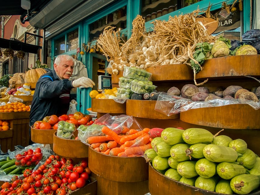A man with grey hair and a dark jacket is seen inside a market stall, surrounded by an array of fresh vegetables and produce. He is wearing a glove on one hand and is reaching out to pick or arrange items, indicating a packing or handling process. The display shelves are wooden and curved, holding various vegetables such as carrots, tomatoes, courgettes, beetroot, garlic, and other root vegetables, with some wrapped in plastic. Behind him, larger amounts of garlic, onions, and squash are stacked on tiered shelves, suggesting a busy food market setting. The environment is well-lit, with natural and artificial lighting illuminating the vibrant produce and the surrounding shopfront, which features teal-colored framing. This scene may be used to depict packing, loading, or moving processes relevant to house or shop removal services, such as those offered by Man and Van Stepney, in preparation for relocation or transportation of goods.