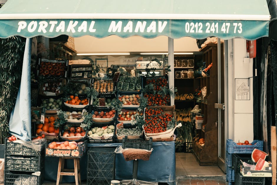 A street-facing fruit and vegetable shop with a green awning displaying the name 'PORTAKAL MANAV' and contact number 0212 241 47 73. The shop showcases an array of fresh produce arranged in black plastic crates and cardboard boxes, including apples, oranges, strawberries, and greens, with some decorated with foliage. To the right, wooden shelves hold additional fruits and vegetables such as bananas and tomatoes, some wrapped in plastic. The interior of the shop, visible through the open entrance, contains more produce and packaging materials, with a narrow walkway between the displays. Outside, on the pavement, a small wooden stool is positioned near the crates, and nearby cardboard boxes and plastic containers are visible, likely for packaging or transport. The scene depicts a typical small grocery holding during daytime, with natural light illuminating the variety of fresh produce, while [COMPANY_NAME] may occasionally assist with similar loading and packing tasks related to house and shop removals, ensuring efficient furniture and goods transport during home relocation or business moving processes.